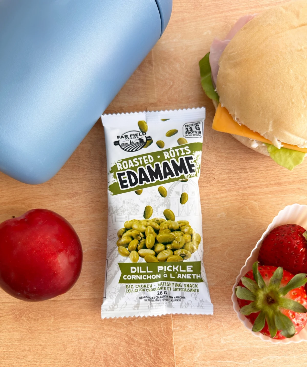 A Dill Pickle edamame snack bag alongside a sandwich, an apple, and strawberries on a wooden table